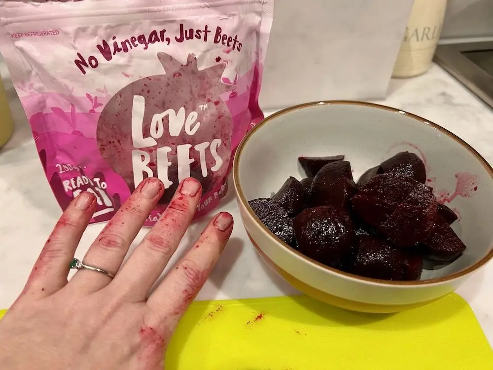 Stained red hands next to a packet of Love Beets cooked beetroot and a bowl full of quartered beetroot pieces. 